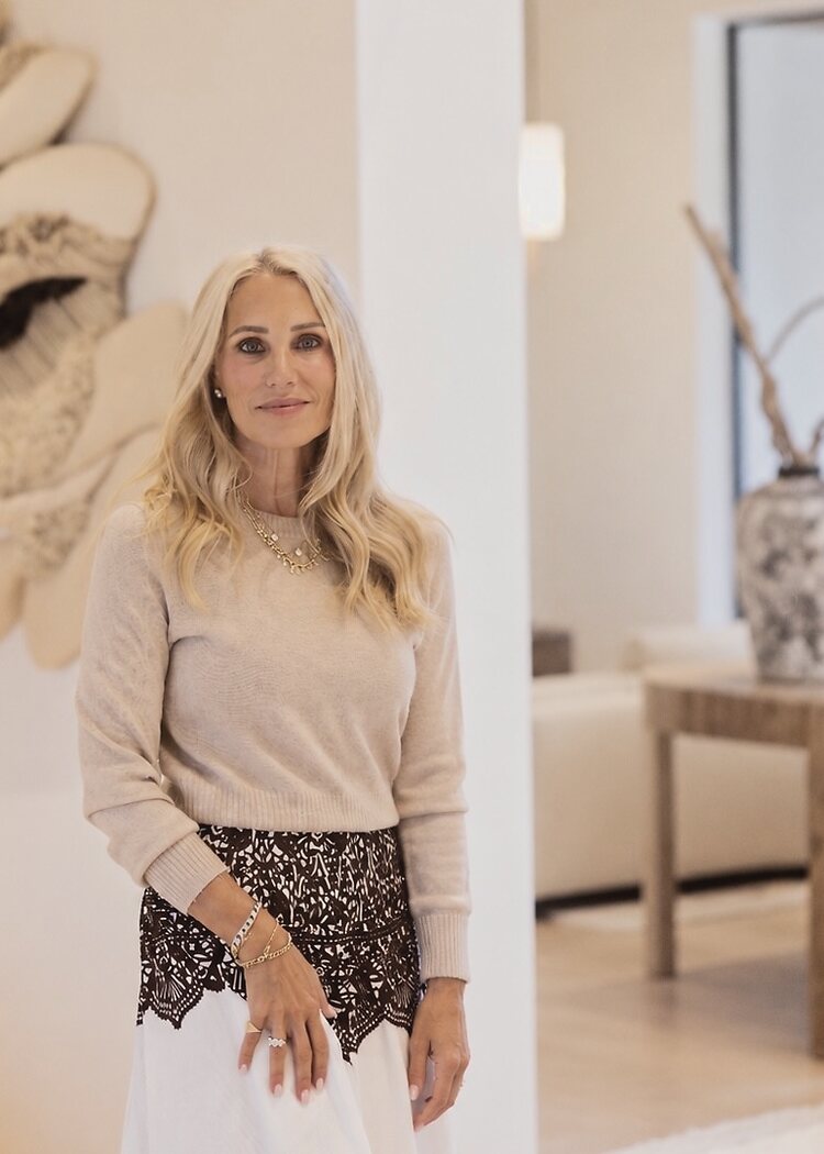 Hailey Lustig standing in a bright, neutral-toned room, wearing a camel sweater, a black lace top and a white skirt